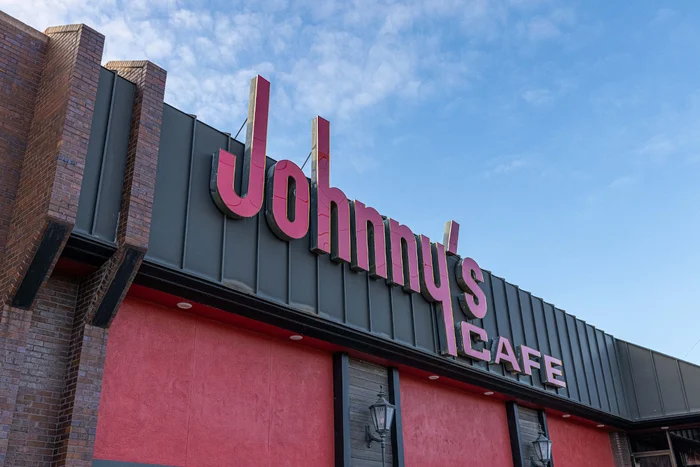 The red script Johnny’s Café sign on the dark metal fascia, against a blue sky.